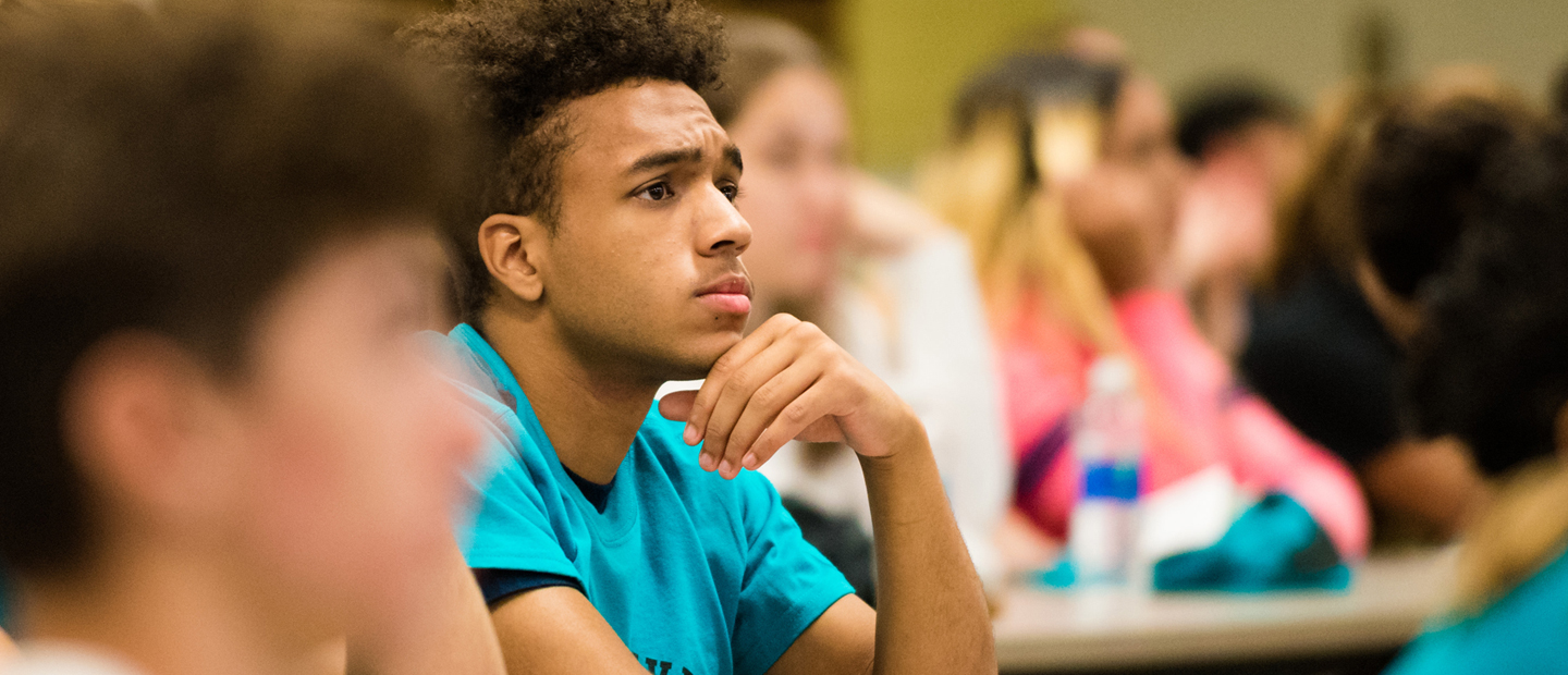 male student in a blue shirt with his chin resting on his hand in a full classroom