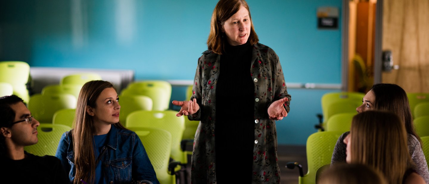 Students sitting at desks look towards a woman who stands speaking