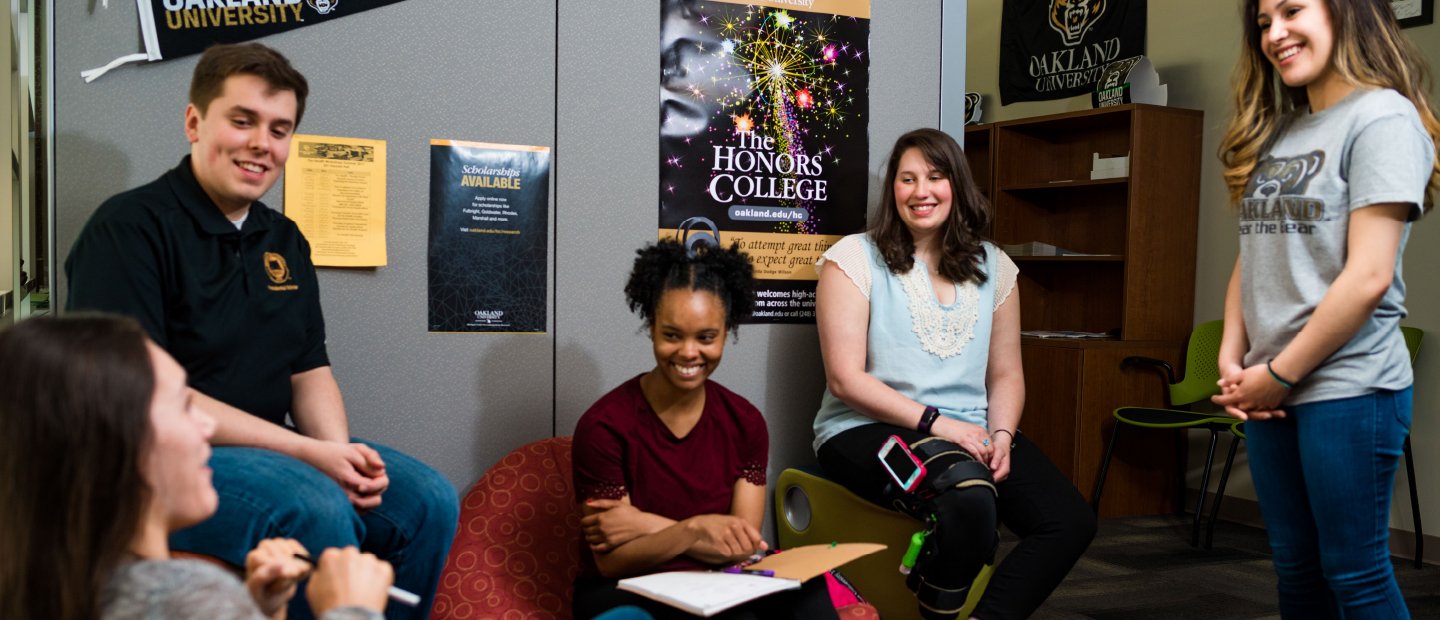 A group of young people sits in an office, talking and laughing. An Honors College poster can be seen in the background.