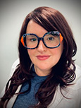 Headshot: Dark haired woman in glasses in blue top and lt. grey jacket smiling at the camera.